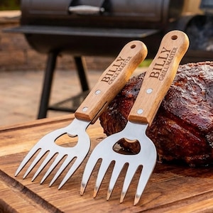 May include: Two stainless steel grilling forks with wooden handles. The handles are engraved with "BILLY GRILL MASTER". The forks are resting on a wooden cutting board next to a large piece of cooked meat.
