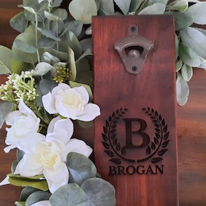 May include: A wooden bottle opener with a dark brown finish. The opener has a metal bottle cap opener and a personalized inscription that reads "B BROGAN" with a laurel wreath design.