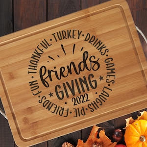May include: A brown bamboo cutting board with silver handles. The board is engraved with the text "Friendsgiving 2022" in a circle with the words "Fun, Thankful, Turkey, Drinks, Games, Laughs, Pie, Friends" around the circle.