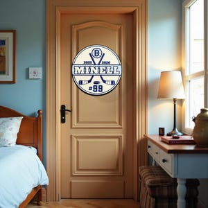 Peut inclure: Un panneau rond, blanc et bleu sur le th&egrave;me du hockey est mont&eacute; sur une porte marron clair. Le panneau affiche le nom "MINELL", le num&eacute;ro "99" et des crosses de hockey crois&eacute;es. Il comprend &eacute;galement les ann&eacute;es "2025" et "2026".