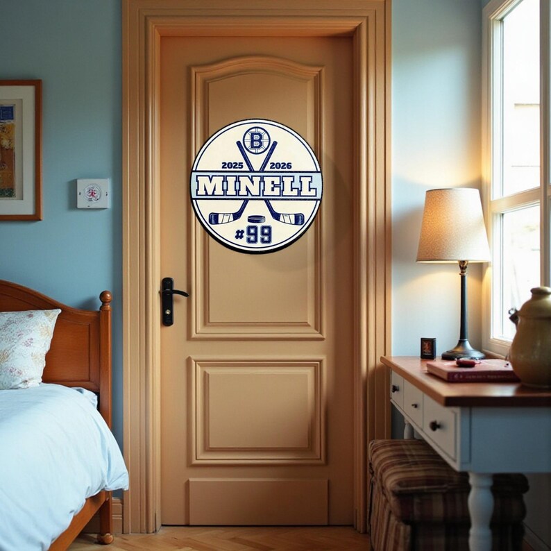 Peut inclure: Un panneau rond, blanc et bleu sur le th&egrave;me du hockey est mont&eacute; sur une porte marron clair. Le panneau affiche le nom "MINELL", le num&eacute;ro "99" et des crosses de hockey crois&eacute;es. Il comprend &eacute;galement les ann&eacute;es "2025" et "2026".