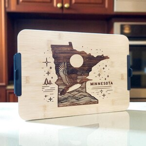 May include: A rectangular wooden serving tray with a laser-etched design of the state of Minnesota. The design includes a loon, a full moon, and the word "MINNESOTA". The tray has black handles on each side.