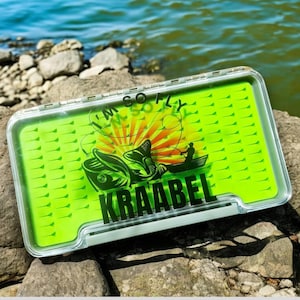 May include: A green fly fishing box with a white lid. The box has a graphic of a fish and the text "I'm So Fly Kraabel".