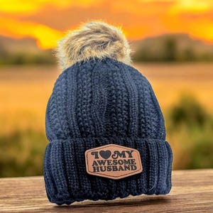 Puede incluir: Un gorro de punto trenzado azul marino oscuro con un pomp&oacute;n de piel sint&eacute;tica en la parte superior. El frente presenta un parche de cuero color canela de forma hexagonal con el texto "I heart MY AWESOME HUSBAND" en negro, con un coraz&oacute;n reemplazando la palabra heart.