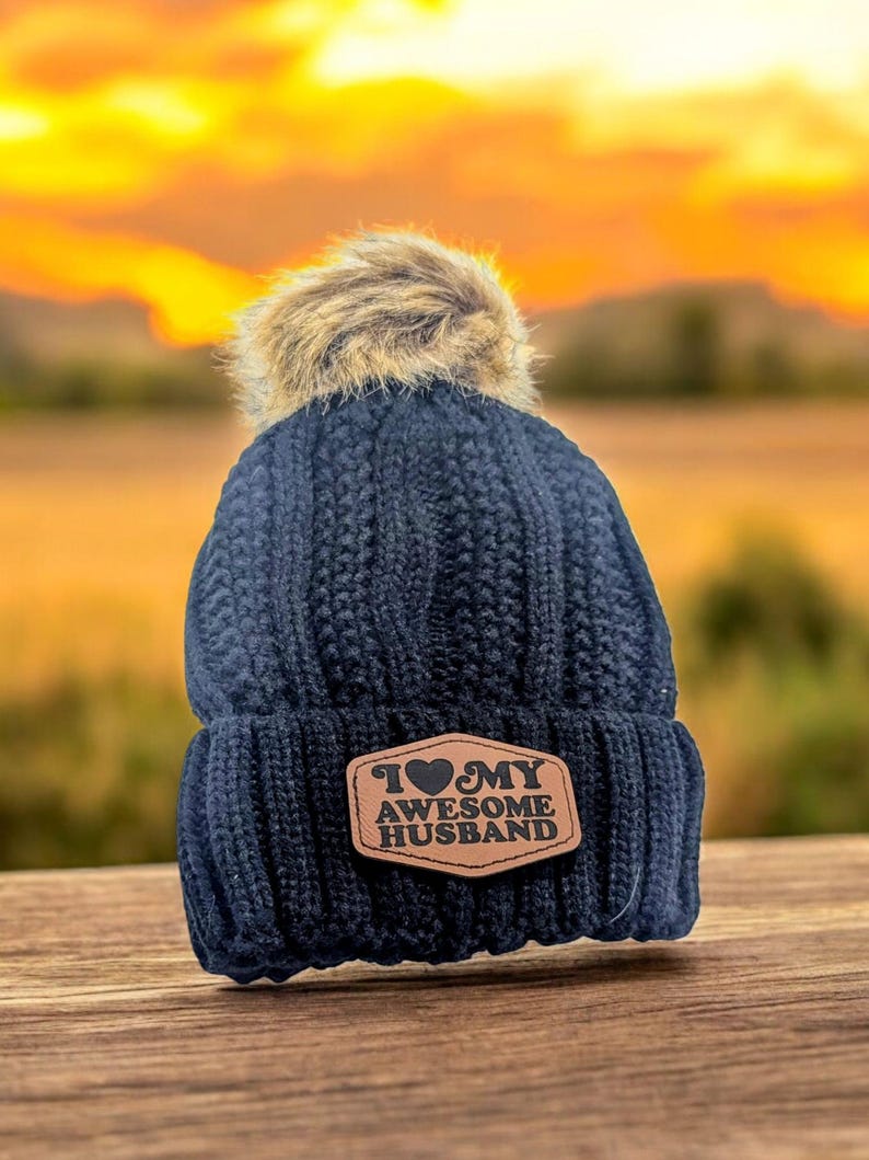 Puede incluir: Un gorro de punto trenzado azul marino oscuro con un pomp&oacute;n de piel sint&eacute;tica en la parte superior. El frente presenta un parche de cuero color canela de forma hexagonal con el texto "I heart MY AWESOME HUSBAND" en negro, con un coraz&oacute;n reemplazando la palabra heart.