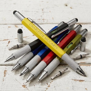 May include: A collection of multi-tool pens in various colors, including yellow, black, blue, red, and green. Each pen has a silver textured grip and a stylus tip. The yellow pen is inscribed with "CHARLOTTE MCCARTY".