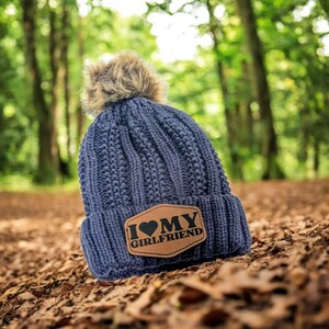 Puede incluir: Un gorro de punto azul marino con un pomp&oacute;n de piel sint&eacute;tica marr&oacute;n. El gorro tiene un parche de cuero marr&oacute;n con el texto "I ❤️ MY GIRLFRIEND". El gorro est&aacute; sobre un lecho de hojas ca&iacute;das.