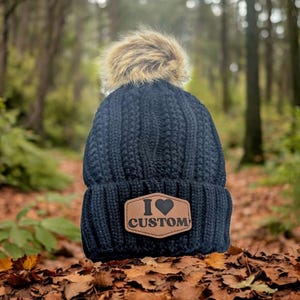 Puede incluir: Un gorro de punto azul marino con un pomp&oacute;n de piel sint&eacute;tica. El gorro presenta un parche de cuero marr&oacute;n con las palabras "I Custom" y un s&iacute;mbolo de coraz&oacute;n. El gorro est&aacute; sobre un lecho de hojas de oto&ntilde;o.
