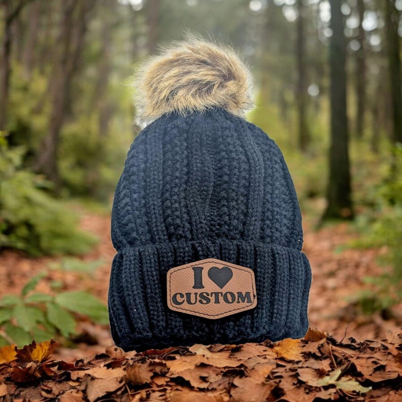 Puede incluir: Un gorro de punto azul marino con un pomp&oacute;n de piel sint&eacute;tica. El gorro presenta un parche de cuero marr&oacute;n con las palabras "I Custom" y un s&iacute;mbolo de coraz&oacute;n. El gorro est&aacute; sobre un lecho de hojas de oto&ntilde;o.