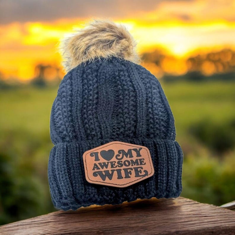 Puede incluir: Un gorro de punto trenzado azul marino oscuro con un pomp&oacute;n de piel sint&eacute;tica beige en la parte superior. Un parche de cuero marr&oacute;n est&aacute; cosido en el frente con el texto I HEART MY AWESOME WIFE, usando un coraz&oacute;n por la palabra heart.