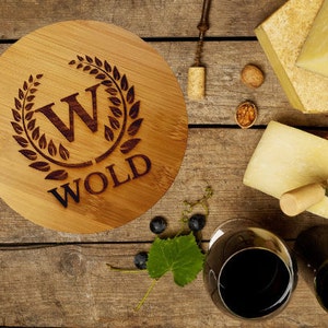 May include: A round wooden cheese board with a personalized engraved design. The design features a wreath with the initials "W" and the name "WOLD".