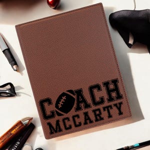 May include: A brown leatherette notebook with a football texture. The cover features the words "COACH MCCARTY" in black block letters with a football graphic. Pens and other items are scattered around the notebook.