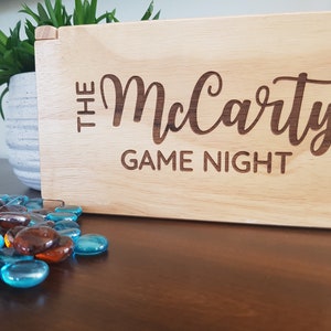 May include: A wooden box with the text "The McCarty Game Night" engraved on the front. The box is sitting on a wooden table with blue and brown glass stones scattered in front of it.