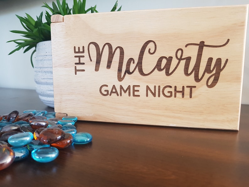 May include: A wooden box with the text "The McCarty Game Night" engraved on the front. The box is sitting on a wooden table with blue and brown glass stones scattered in front of it.
