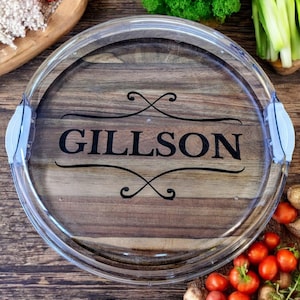 May include: A round wooden serving tray with a clear plastic rim and handles. The tray has a wood grain pattern and the name "Gillson" is engraved in black lettering.