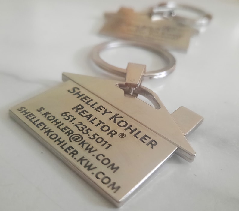 Op de afbeelding: Zilverkleurige sleutelhanger in de vorm van een huis met de tekst "Shelley Kohler, Realtor" en contactgegevens. De sleutelhanger is bevestigd aan een zilveren sleutelring. Een andere sleutelhanger is enigszins onscherp op de achtergrond.