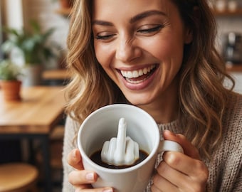 May include: A white ceramic coffee mug with a handle, filled with dark liquid and a white, hand-shaped sculpture inside, giving the middle finger. The mug is held by a person in a beige sweater.