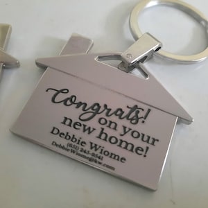 Op de afbeelding: Zilverkleurige sleutelhanger in de vorm van een huis met een sleutelring. De sleutelhanger heeft de woorden "Congrats on your new home!" gegraveerd, samen met een naam en contactgegevens.