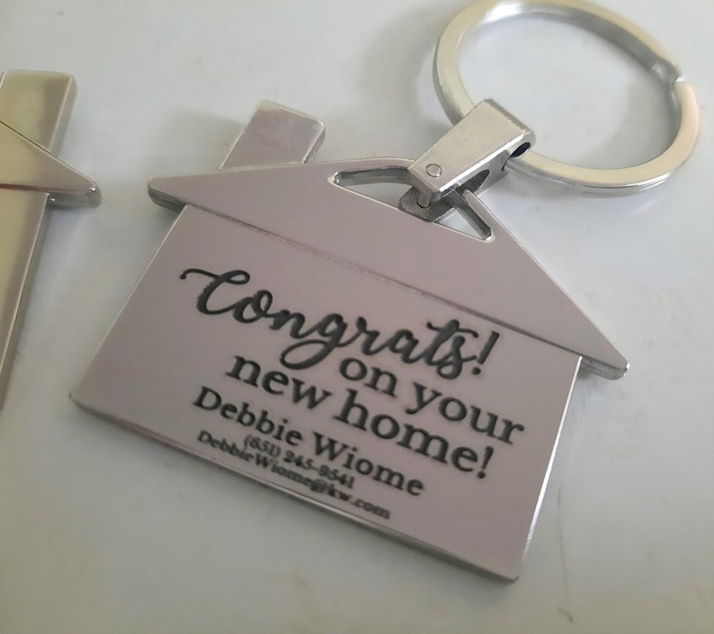 Op de afbeelding: Zilverkleurige sleutelhanger in de vorm van een huis met een sleutelring. De sleutelhanger heeft de woorden "Congrats on your new home!" gegraveerd, samen met een naam en contactgegevens.
