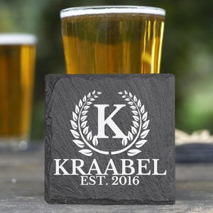 May include: Black slate coaster with a white wreath and the letter K in the center. The text "Kraabel Est 2016" is below the wreath.