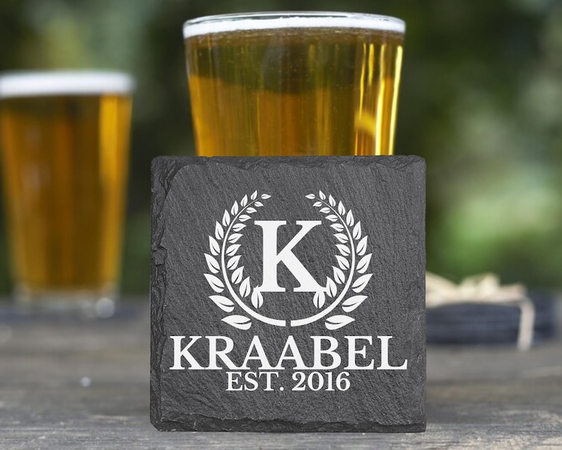 May include: Black slate coaster with a white wreath and the letter K in the center. The text "Kraabel Est 2016" is below the wreath.