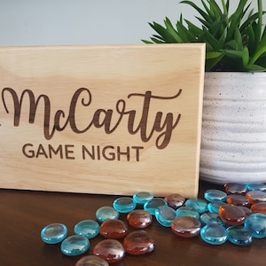 May include: A wooden sign with the text "The McCarty Game Night" engraved on it. The sign is sitting on a wooden table with blue and brown glass stones scattered around it.