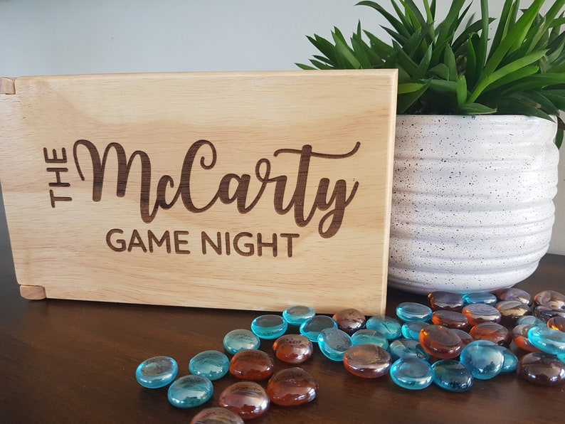 May include: A wooden sign with the text "The McCarty Game Night" engraved on it. The sign is sitting on a wooden table with blue and brown glass stones scattered around it.