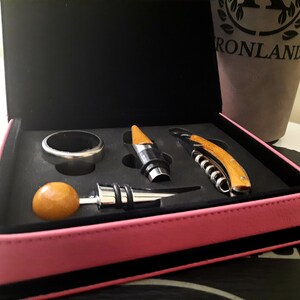May include: A pink leatherette case with a black interior holds a set of wine accessories. The set includes a bottle opener, a cork screw, a bottle stopper, and a ring for securing a bottle.
