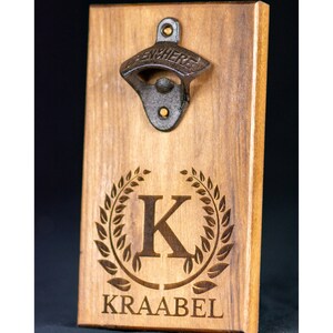 May include: Wooden bottle opener with a metal opener and a personalized engraved design. The design features a wreath with the letter K and the name "KRAABEL" below.