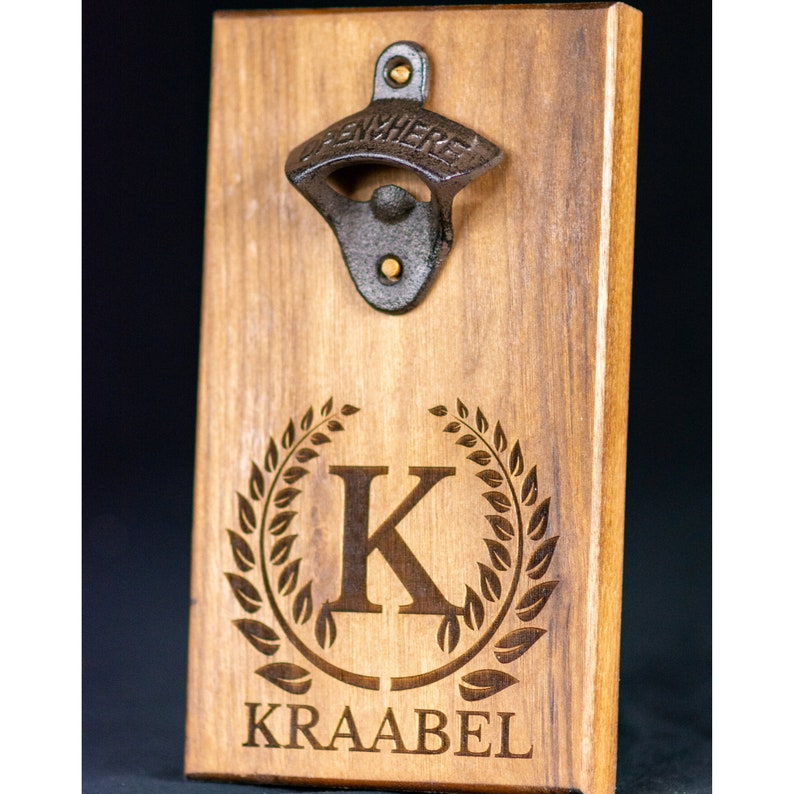May include: Wooden bottle opener with a metal opener and a personalized engraved design. The design features a wreath with the letter K and the name "KRAABEL" below.