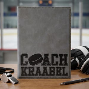 Op de afbeelding: Een grijze notitieboek met de tekst "COACH KRAABEL" en een hockey puck afbeelding. Een zilveren fluitje, zwarte pen en hockeyhandschoenen liggen op een houten oppervlak. Het notitieboek is waarschijnlijk voor een hockeycoach.