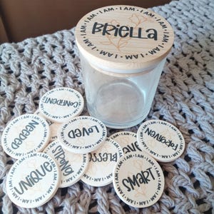 Op de afbeelding: Een heldere glazen pot met een houten deksel, met de naam "Briella" en bloemmotieven. Rondom de pot liggen verschillende witte ronde tokens met bemoedigende woorden als "Geliefd" en "Slim" in zwarte tekst. De woorden "IK BEN" zijn gedrukt.