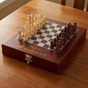 May include: A wooden chess set with a dark brown case and a checkered board. The chess pieces are arranged on the board, with light and dark pieces facing each other. The case has a gold logo and the text "Thank You - ADAMS REALTY".