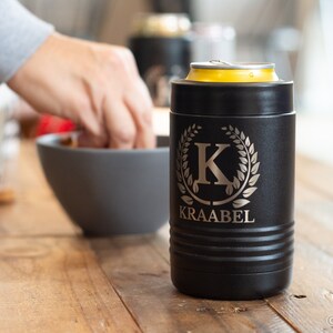 May include: Black insulated can cooler with a silver monogram of the letter "K" surrounded by a laurel wreath. The text "KRAABEL" is engraved below the monogram.