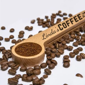 May include: A wooden coffee scoop with the text "Linda's Coffee" engraved on it. The scoop is filled with ground coffee and surrounded by coffee beans.