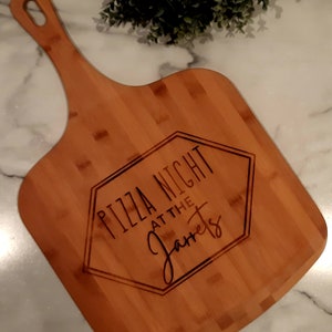 以下が含まれることがあります： ハンドル付きの木製のピザサービングボード。ボードには「Pizza Night at the Jarrets」というテキストが刻まれています。