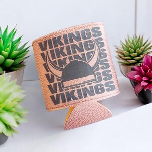 May include: A tan can cooler with the word "VIKINGS" printed in black, along with a Viking helmet graphic. The cooler has a textured surface and is designed to hold a standard-sized beverage can. The background includes potted succulents.