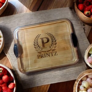 May include: A rectangular bamboo serving tray with a clear lid and black handles. The tray features an engraved design with the letter "P" inside a laurel wreath and the word "PRINTZ" below.