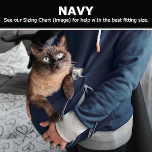 May include: A navy blue hoodie with a pocket for a cat. The cat is a brown tabby with blue eyes. The hoodie has a gray zipper and a gray drawstring.