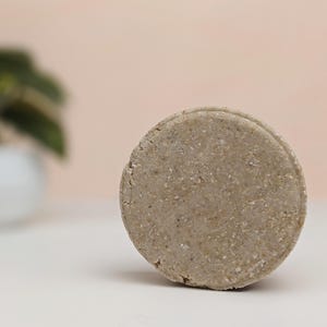 Peut inclure: Un pain de shampooing rond et texturé de couleur grise est au centre de l'image. Il est posé sur une surface blanche, avec un arrière-plan flou présentant une plante en pot. L'esthétique générale est minimaliste et naturelle.