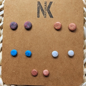 May include: A set of five pairs of round stud earrings displayed on a brown cardboard card with the text NATUKARI. The earrings feature various textures and colours, including dark red, copper, bright blue with leaf patterns, and speckled white.