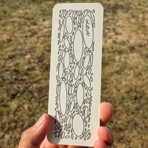 May include: A rectangular bookmark with rounded corners. The bookmark has a black ink design of various sized ovals and small leaf-like shapes. The word "love" is written in cursive. The bookmark is held in a hand against a blurred outdoor background.
