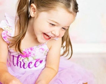 May include: A young girl with blonde hair wearing a purple dress with a floral pattern and the word "three" on the front. She is smiling and looking down. She is wearing a pink flower hair clip.
