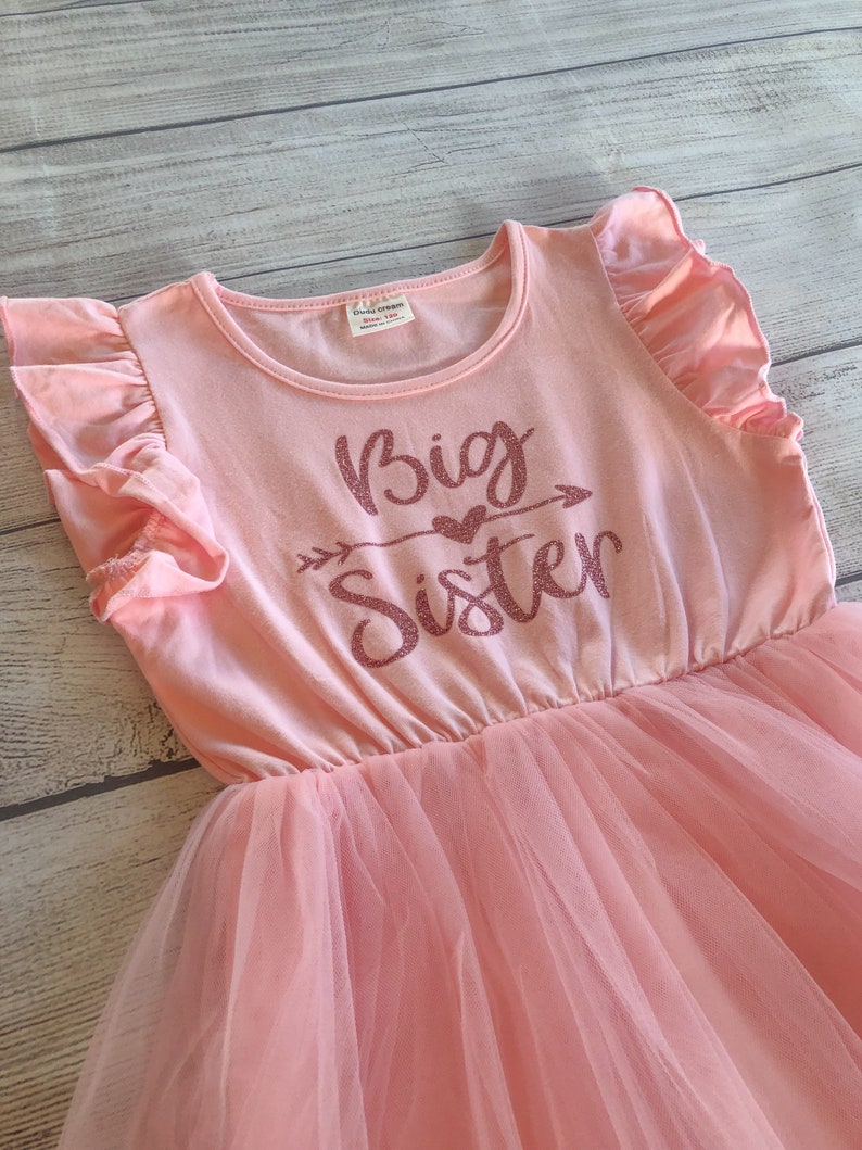 May include: Pink tulle dress with a pink short-sleeved shirt overlay. The shirt has a glittery pink design that says "Big Sister" with an arrow and a heart.