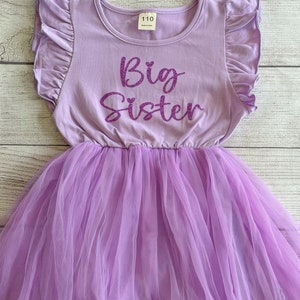 May include: A lavender tulle dress with a purple glitter "Big Sister" graphic on the bodice. The dress has short sleeves with ruffles and a round neckline.