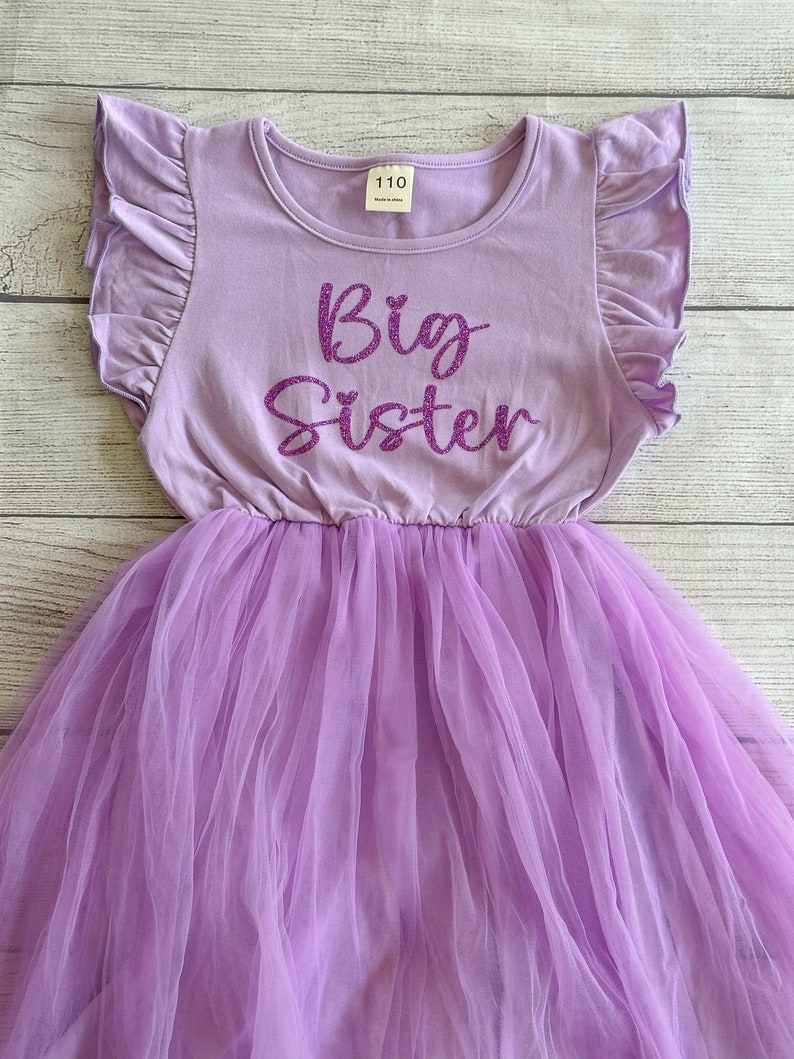 May include: A lavender tulle dress with a purple glitter "Big Sister" graphic on the bodice. The dress has short sleeves with ruffles and a round neckline.