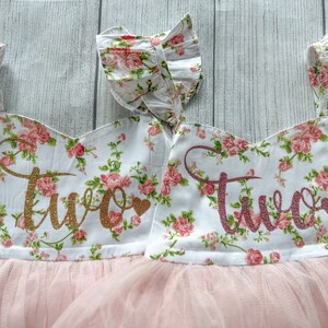May include: Two pink and white floral print dresses with tulle skirts. The dresses have gold glitter "Two" on the front.