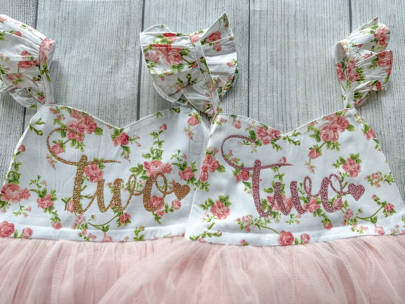 May include: Two pink and white floral print dresses with tulle skirts. The dresses have gold glitter "Two" on the front.