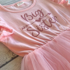 May include: Pink tulle dress with a pink cotton top. The top has the words "Big Sister" in sparkly pink letters with an arrow and a heart.