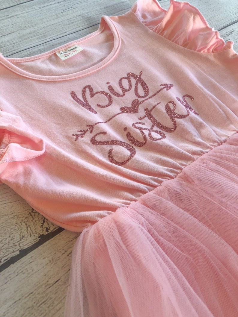 May include: Pink tulle dress with a pink cotton top. The top has the words "Big Sister" in sparkly pink letters with an arrow and a heart.